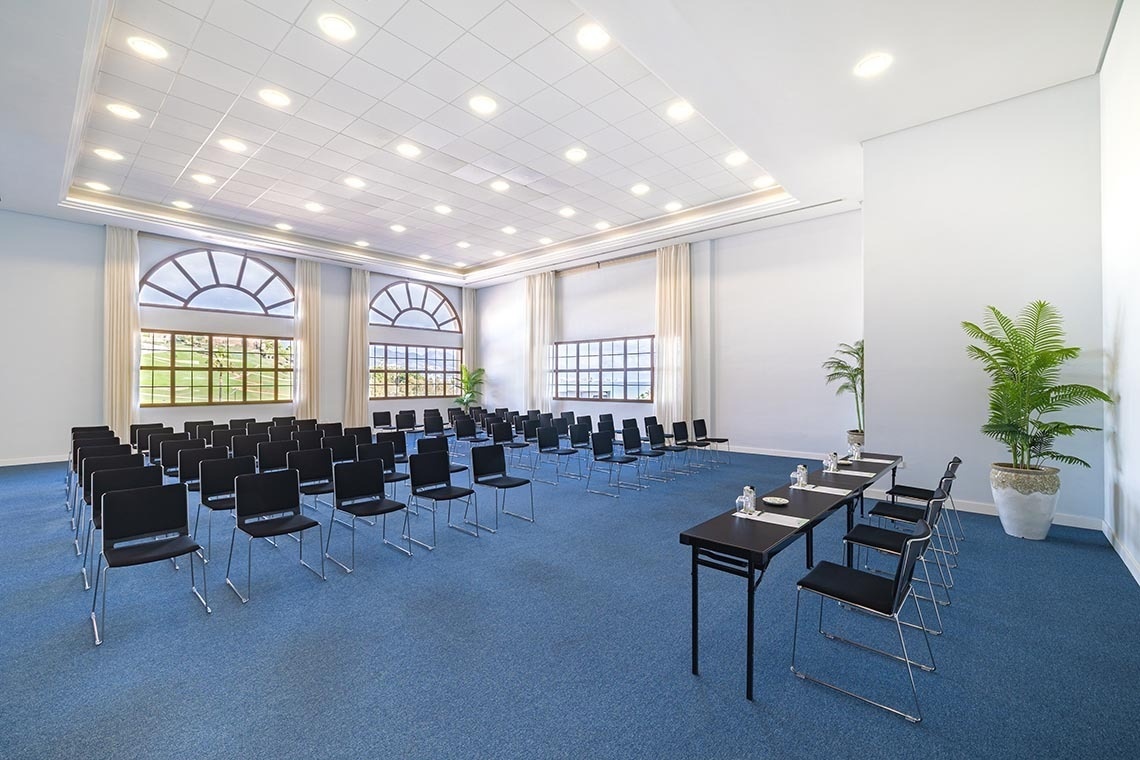 A spacious conference room features rows of black chairs on blue carpet, long tables with chairs, large arched windows revealing outdoor greenery, and potted plants.
