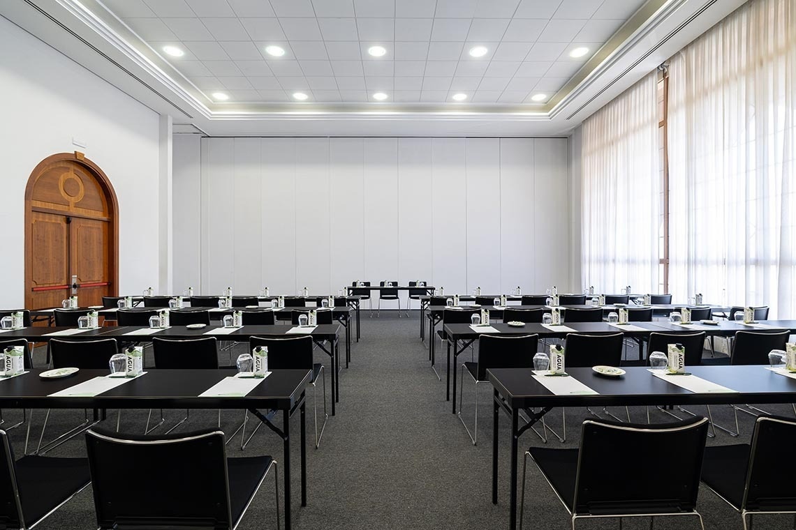 A large, well-lit conference room is set up with multiple rows of black tables and chairs, each prepared with water and materials, featuring a decorative arched wooden door on the left, a modular white wall at the back, and floor-to-ceiling windows with sheer white curtains on the right.