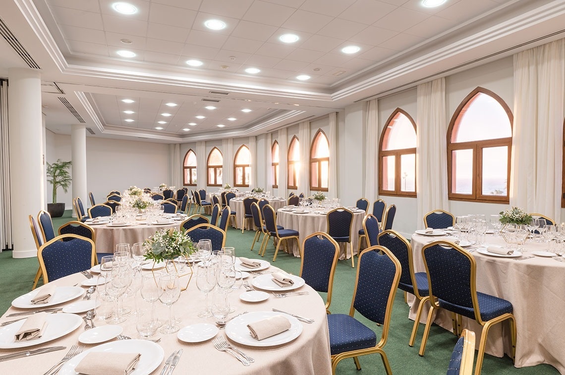 Une grande salle de réception élégante est aménagée avec de nombreuses tables rondes dressées pour un événement, entourées de chaises bleues et illuminée par des fenêtres arquées.