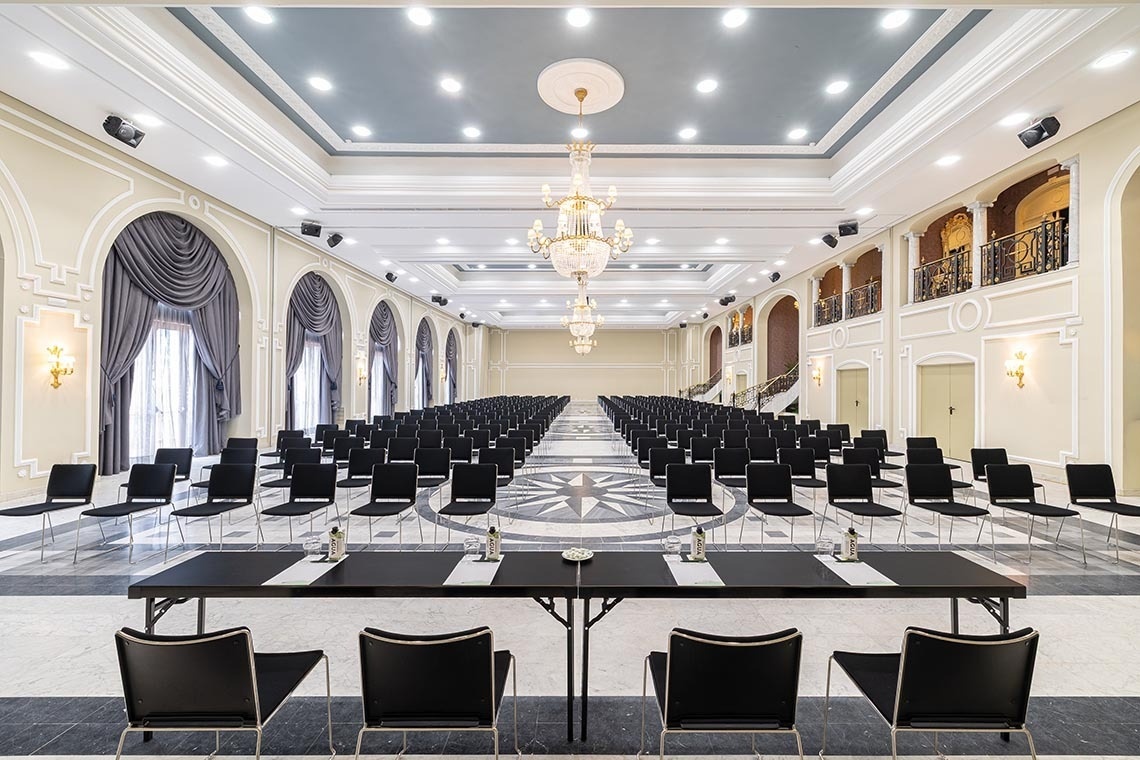 An empty, brightly lit conference room features rows of black chairs on a blue carpet, several side tables, and large arched windows offering views of a green landscape.