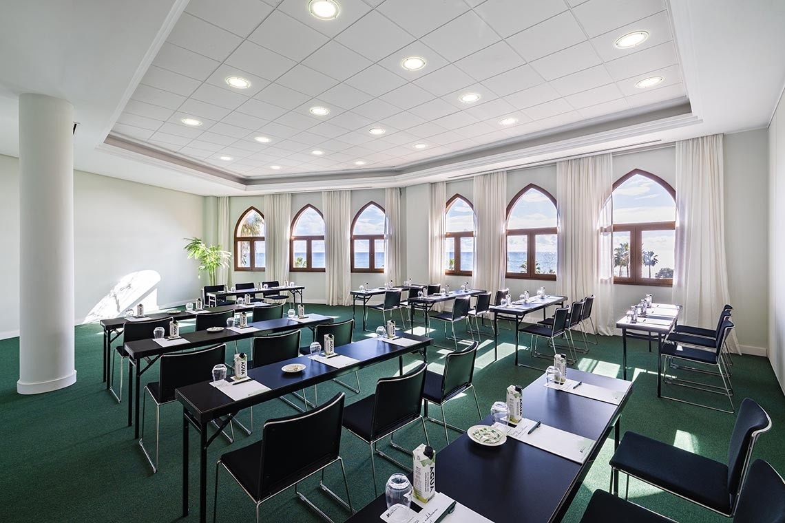A brightly lit conference room is set up with tables and chairs, featuring arched windows that offer a scenic view of the ocean and palm trees.