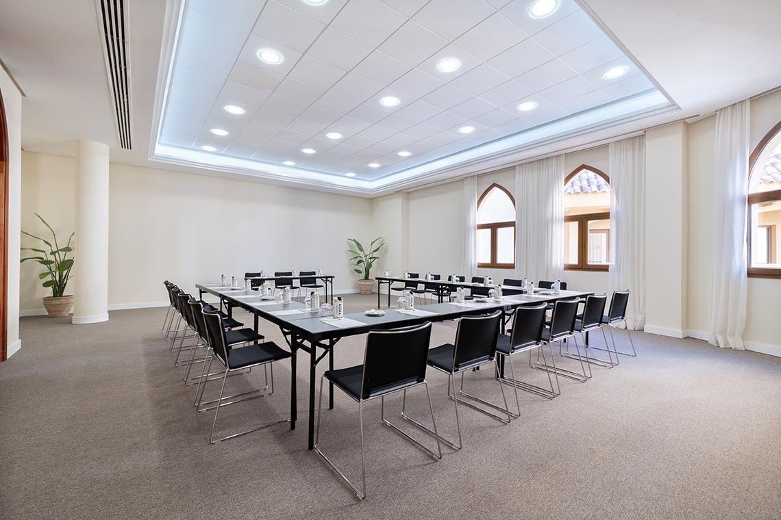 This bright meeting room features a U-shaped table setup with black chairs, a suspended ceiling with recessed lighting and a perimeter light strip, and several arched windows with white curtains along one wall.