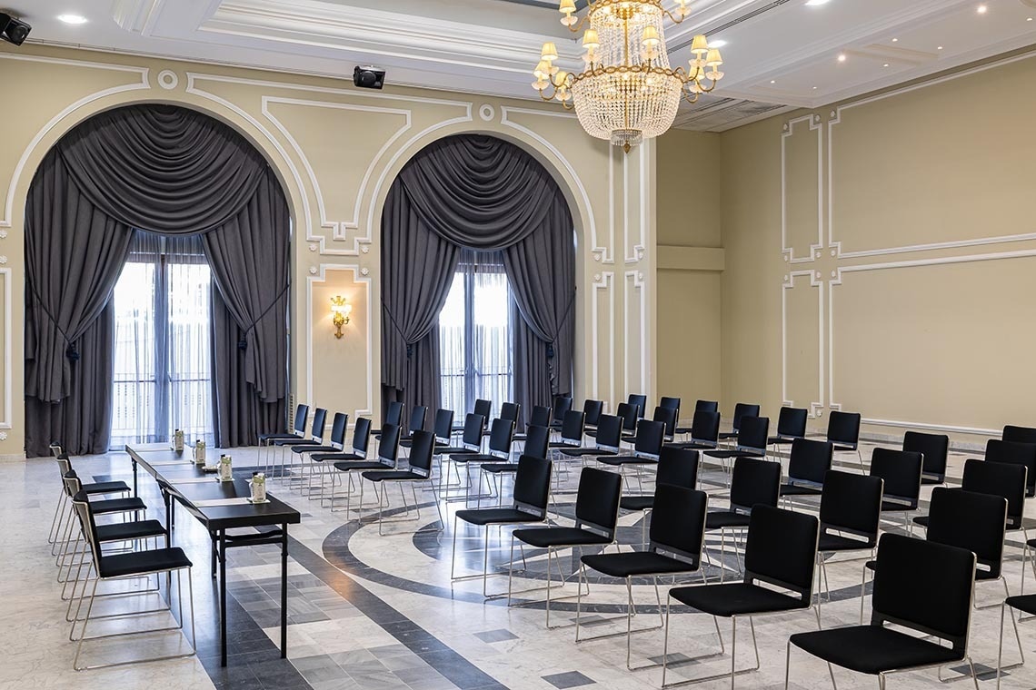 An elegant conference room is set up with multiple rows of black chairs facing a long central table, featuring ornate light yellow walls with large arched windows draped in grey, and illuminated by a grand chandelier and wall sconces.