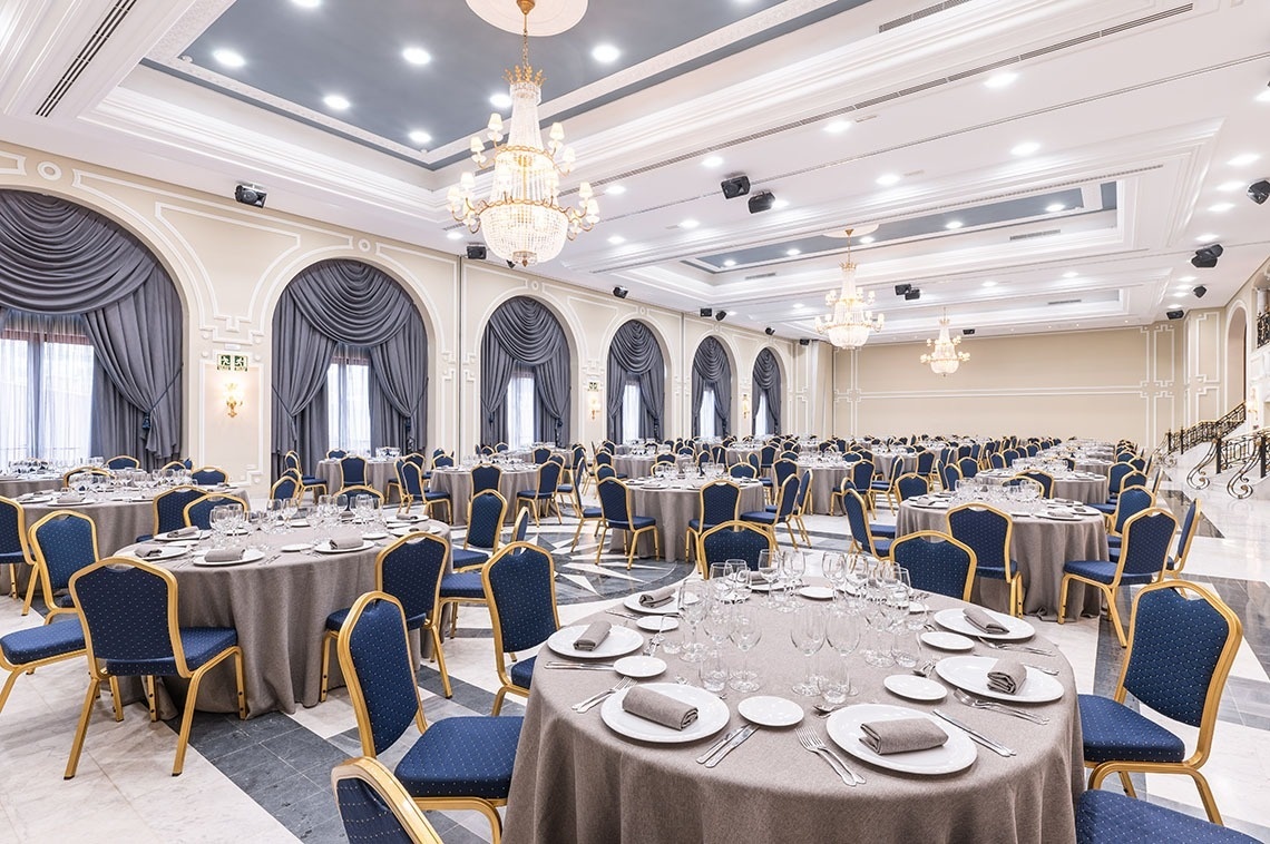 Une vaste et élégante salle de banquet est aménagée avec de nombreuses tables rondes dressées et des chaises bleues et or, sous de grands lustres et le long de fenêtres arquées drapées.