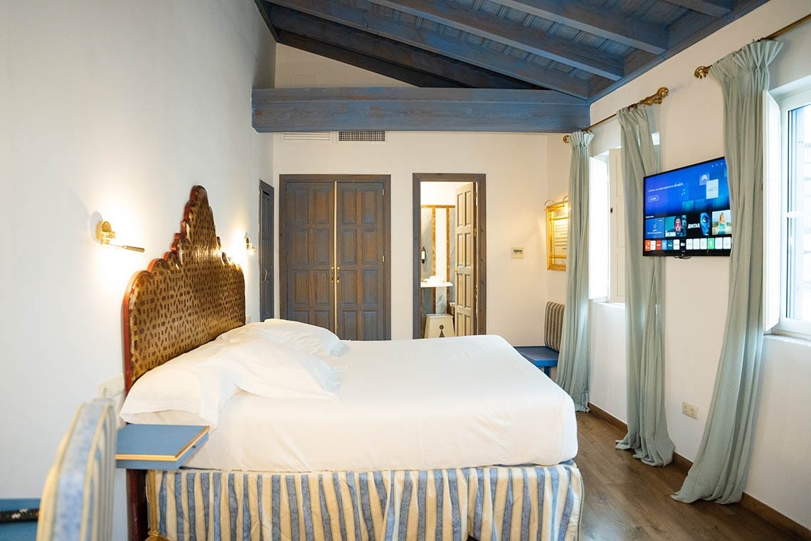 Un dormitorio cálidamente iluminado exhibe una cama grande con un cabecero ornamentado, un techo con vigas azules y detalles en tonos azules como cortinas, una falda de cama a rayas y puertas de madera rústicas.