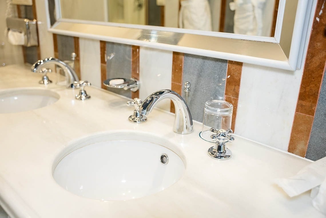 La imagen muestra un tocador de baño con doble lavabo, grifos cromados, una encimera de mármol y una pared de azulejos de mármol a rayas, con un espejo en la parte superior.