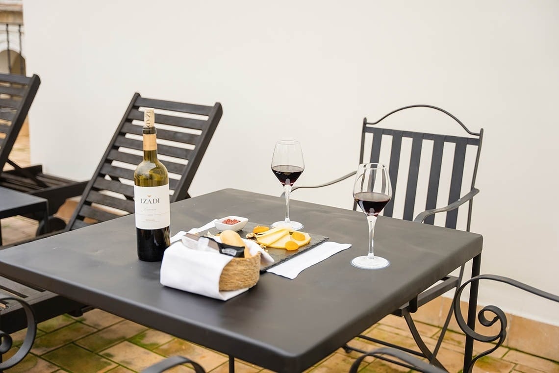 Una mesa exterior cuenta con una botella de vino tinto Izadi, dos copas de vino, queso y pan, con tumbonas y sillas oscuras al fondo.