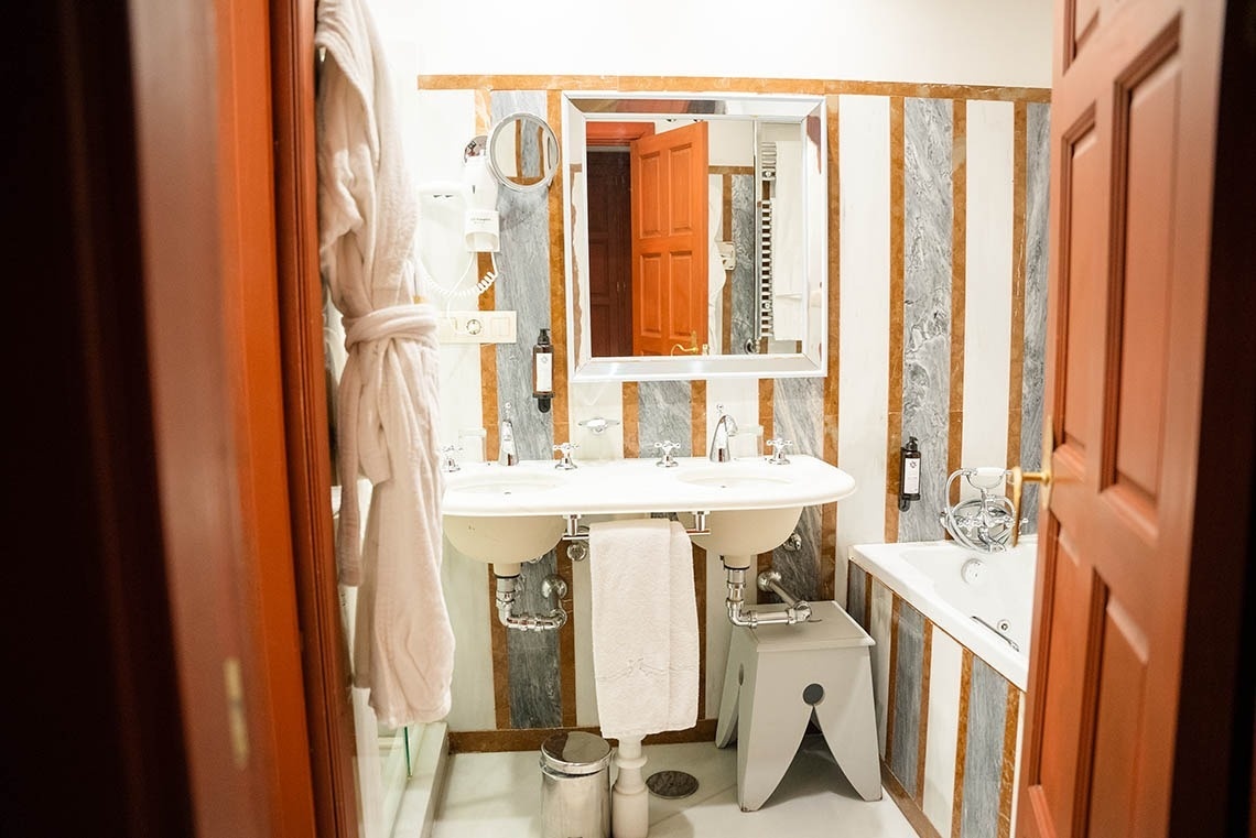 Un cuarto de baño bien iluminado presenta un lavabo doble, un espejo rectangular y una bañera, todo ello enmarcado por paredes a rayas de mármol y una puerta de madera abierta en primer plano.