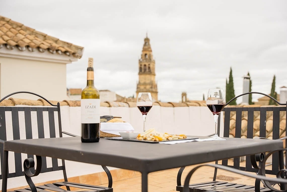 Una mesa con vino y aperitivos se encuentra en una azotea con vistas a una ciudad, destacando una torre campanario en el fondo.