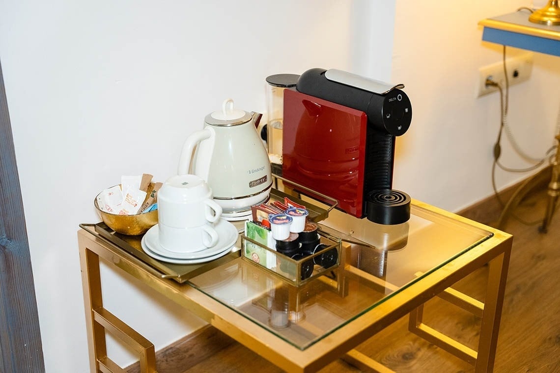 Una estación de café y té con una cafetera roja, un hervidor blanco, tazas y varios suministros está dispuesta sobre una mesa auxiliar de cristal con estructura dorada.