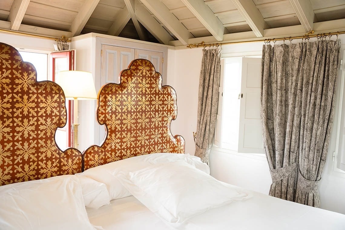 Una cama con un cabecero ornamentado en dorado y marrón se encuentra en una habitación luminosa con techo de vigas de madera y ventanas con cortinas estampadas.