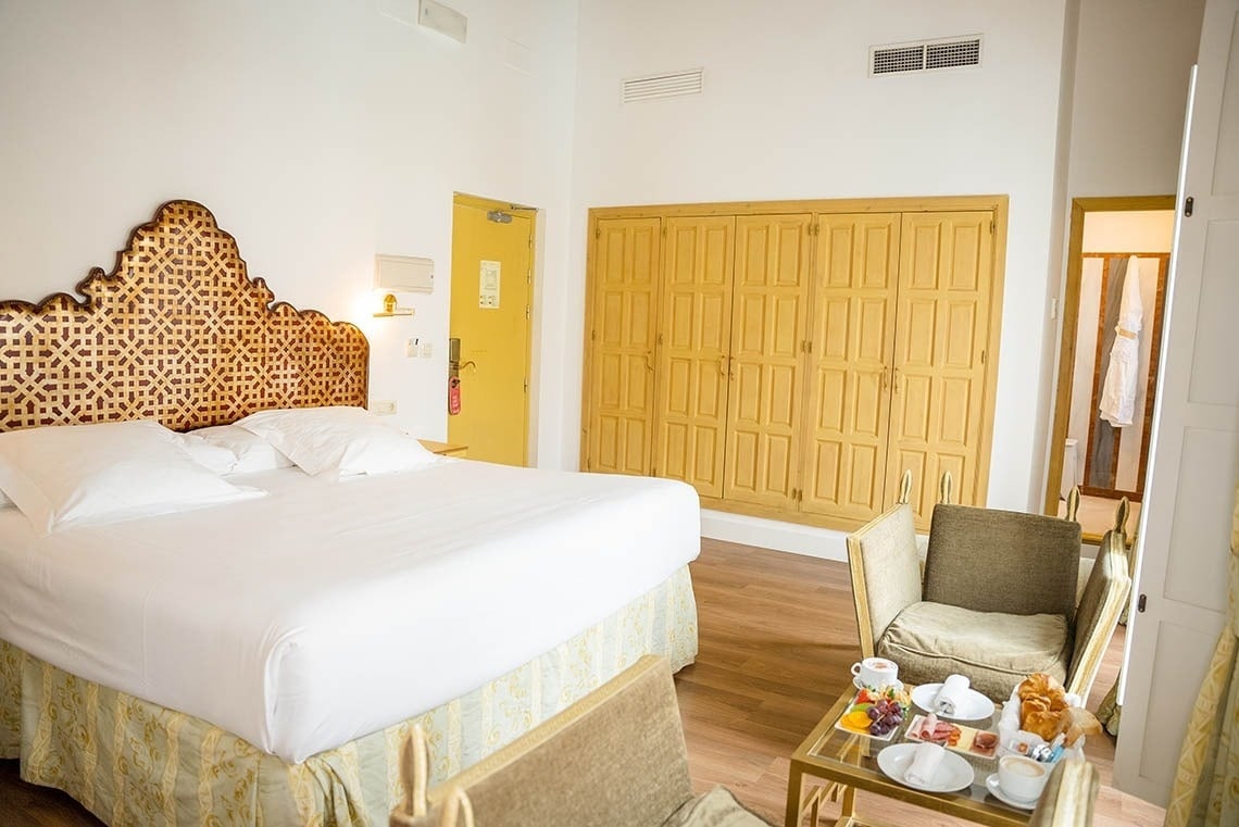 Una luminosa habitación de hotel con una cama grande de cabecero ornamentado, un armario de madera, una puerta amarilla y una mesa con desayuno.