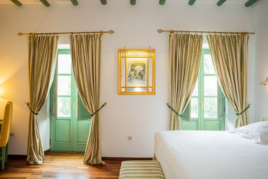 Un dormitorio luminoso con dos ventanas de marcos verdes y cortinas a rayas, un cuadro dorado en el centro y una cama a la derecha.