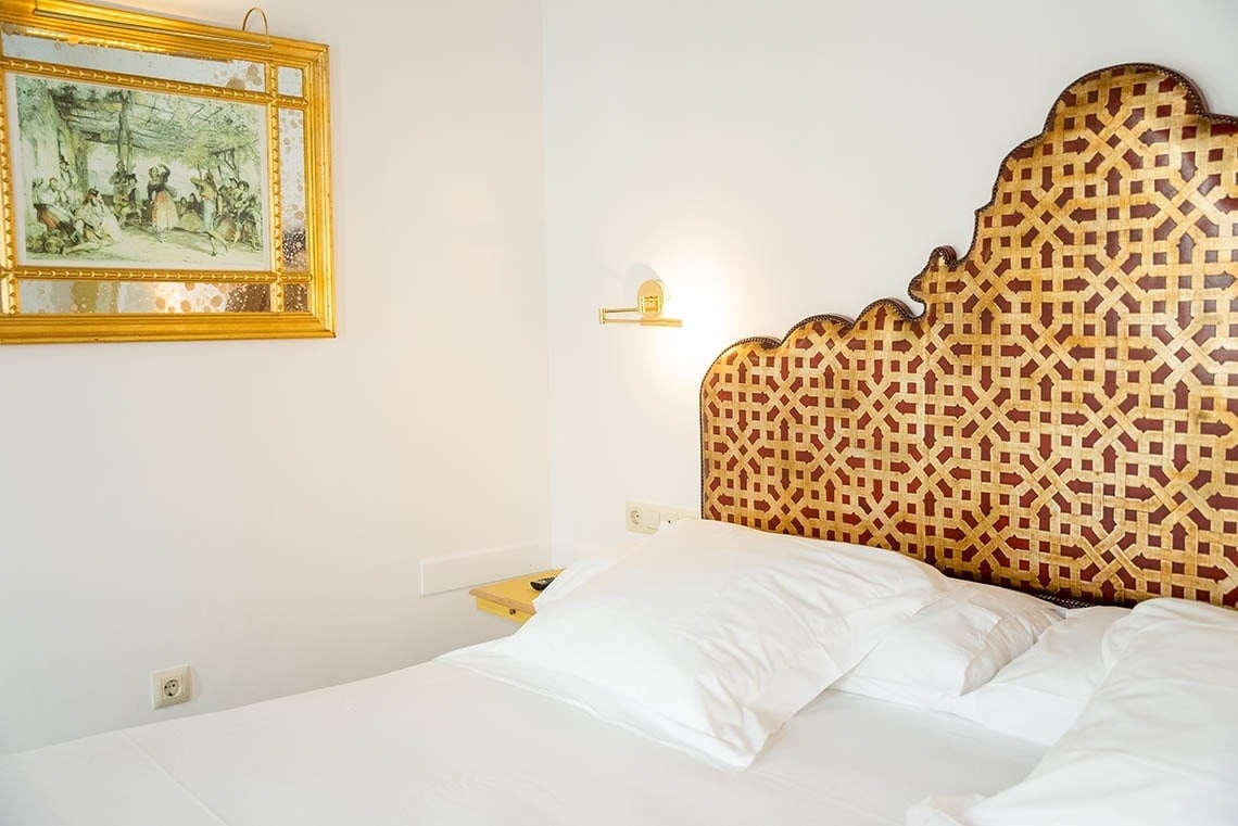Una cama con un llamativo cabecero dorado y rojizo de patrón geométrico, vestida con ropa de cama blanca y con un espejo dorado que enmarca una pintura en la pared.