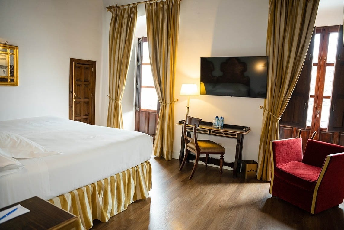 Una habitación de hotel de estilo clásico con una cama, un escritorio con televisor, un sillón rojo y ventanas con contraventanas de madera y cortinas doradas.