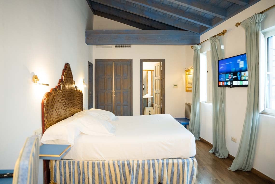 Elegant hotel room in Juderia: Cordoba, featuring a wrought iron bed, blue beamed ceiling, and modern TV.