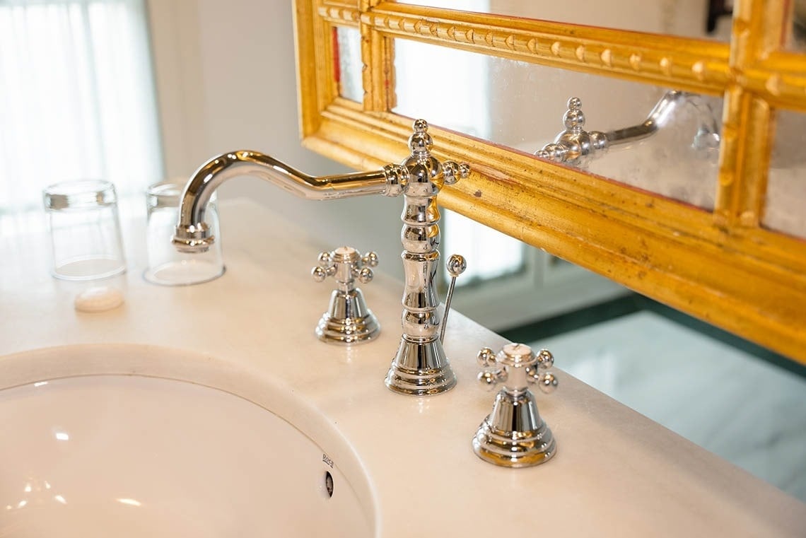 Un primer plano de un lavabo de baño de mármol con grifería cromada, dos vasos, una pastilla de jabón y un espejo con marco dorado.