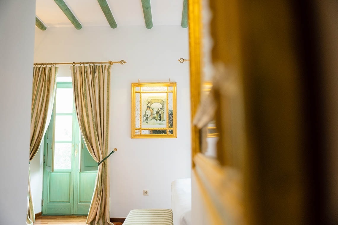 Una habitación luminosa presenta paredes blancas, vigas de techo y puertas de madera en tono verde menta, adornada con cortinas a rayas y un espejo con marco dorado colgado en la pared.