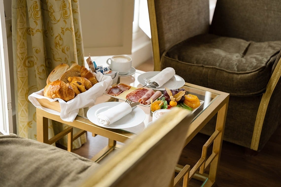 Un desayuno variado, con pan, embutidos, fruta y café, está dispuesto sobre una mesa de cristal entre dos sillones, con una cortina estampada de fondo.