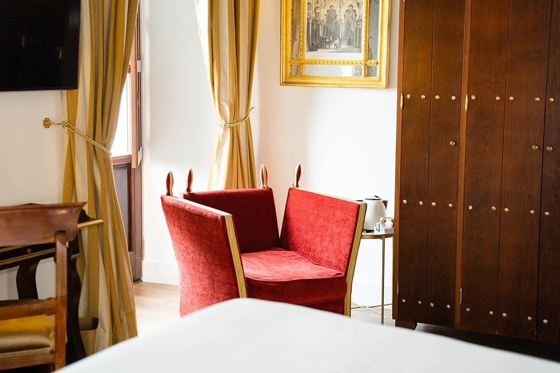Una estancia luminosa decorada con una silla roja de terciopelo, cortinas de rayas doradas y un armario alto de madera oscura con detalles en latón, junto a una pintura enmarcada.