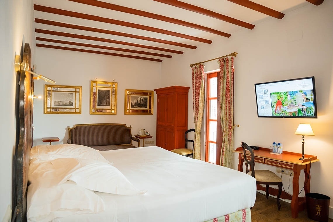 Dormitorio de estilo clásico con techo de vigas de madera vistas, una cama grande, televisión y mobiliario tradicional en tonos cálidos.