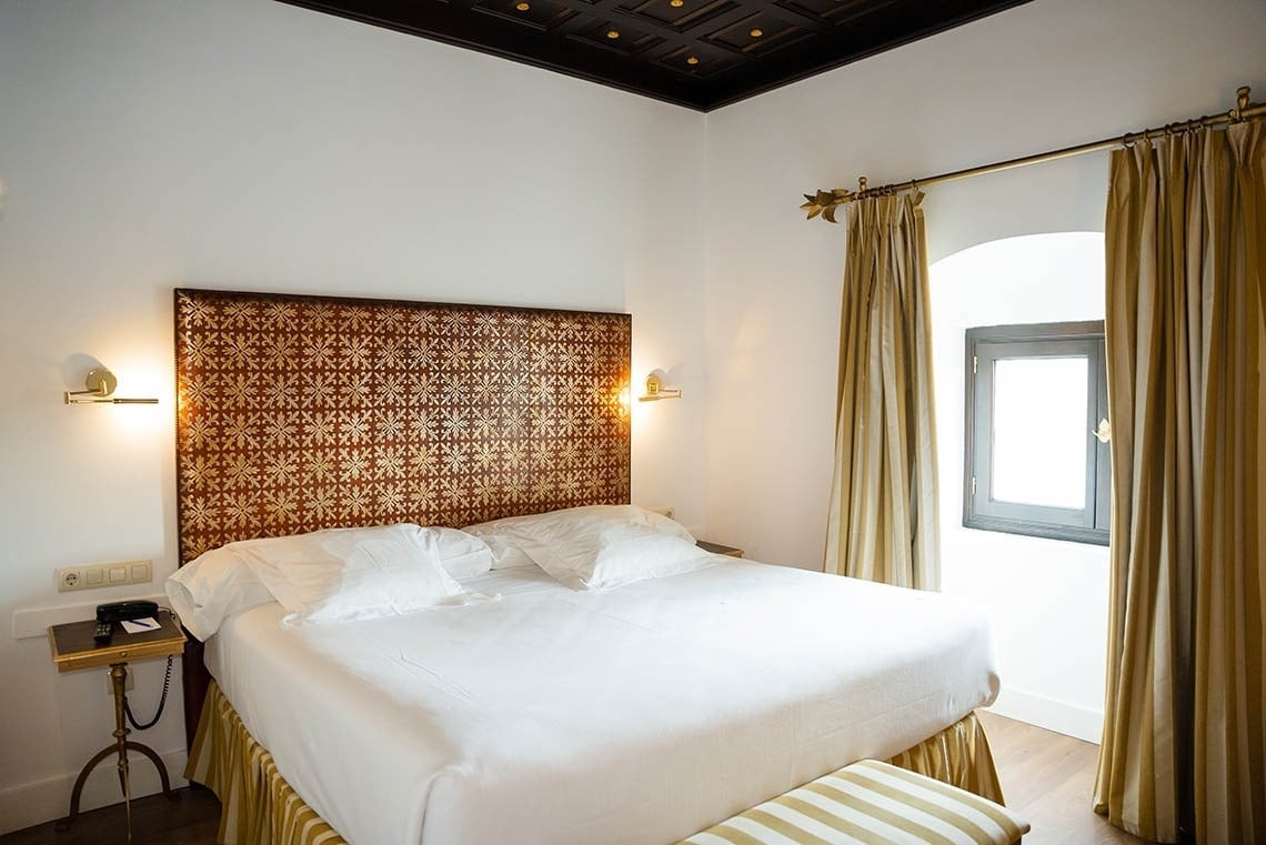 Una habitación de hotel con una cama grande y un cabecero estampado dorado, paredes blancas, cortinas doradas y un techo de madera oscuro.