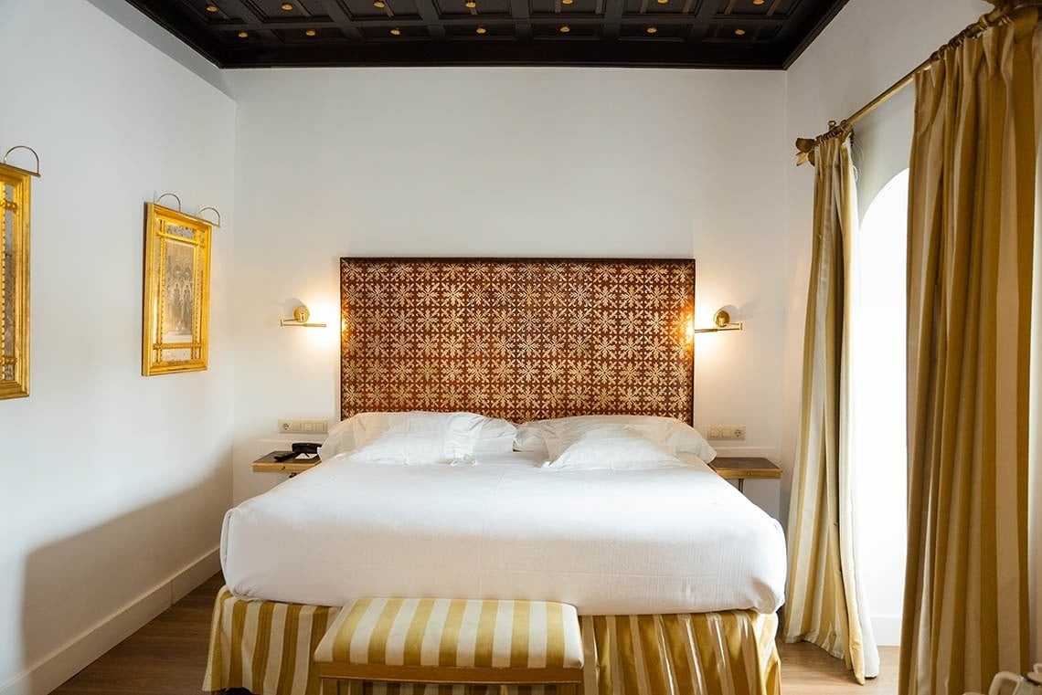 Un dormitorio de hotel elegantemente decorado con una cama grande, un cabecero estampado en tonos dorados y marrones, paredes blancas, un techo artesonado oscuro y cortinas doradas a rayas.