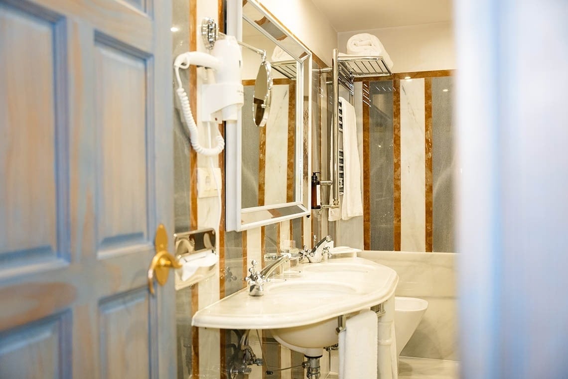 Un elegante baño con paredes de mármol, lavabo doble, espejo y secador de pelo es visible a través de una puerta de madera abierta.