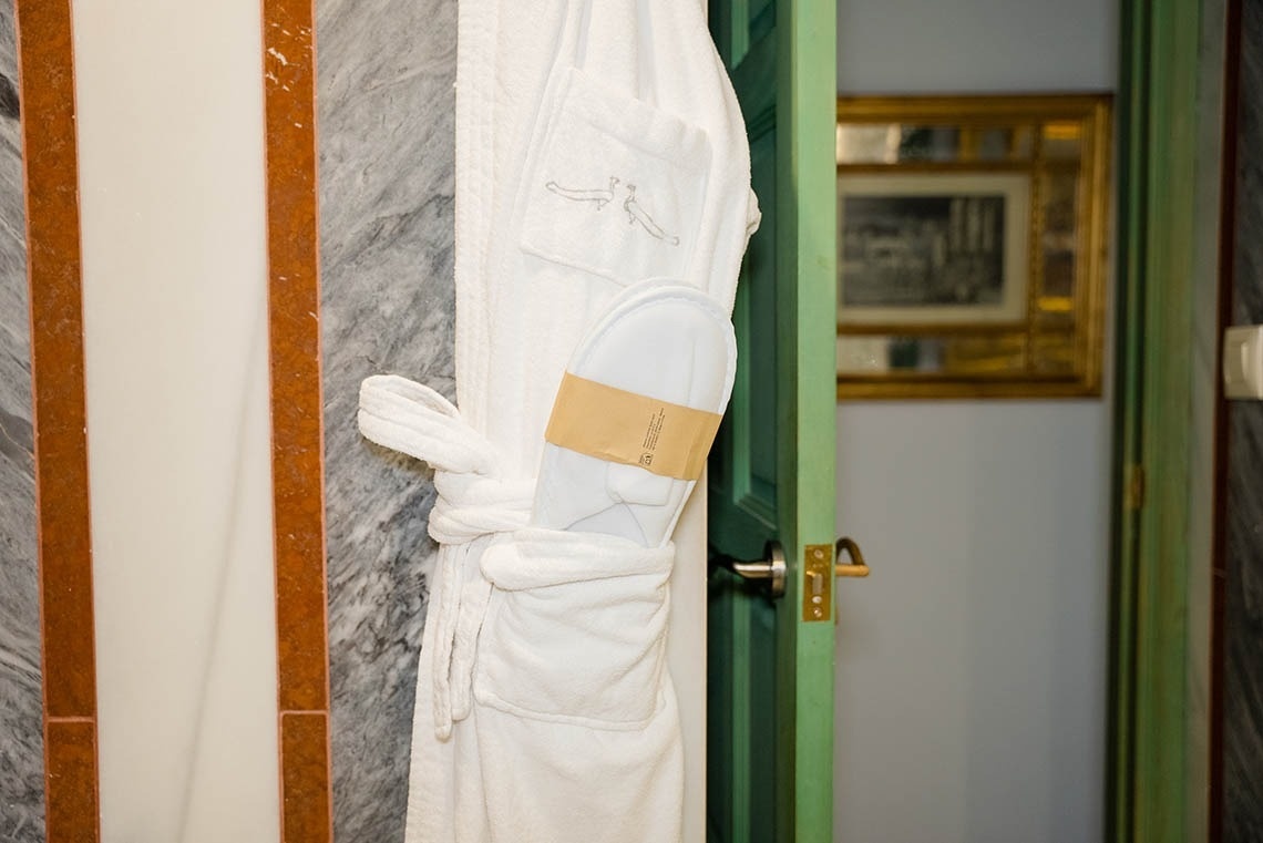 Un albornoz blanco con zapatillas en el bolsillo cuelga junto a una pared de mármol gris con franjas naranjas, con una puerta verde entreabierta que muestra un cuadro al fondo.