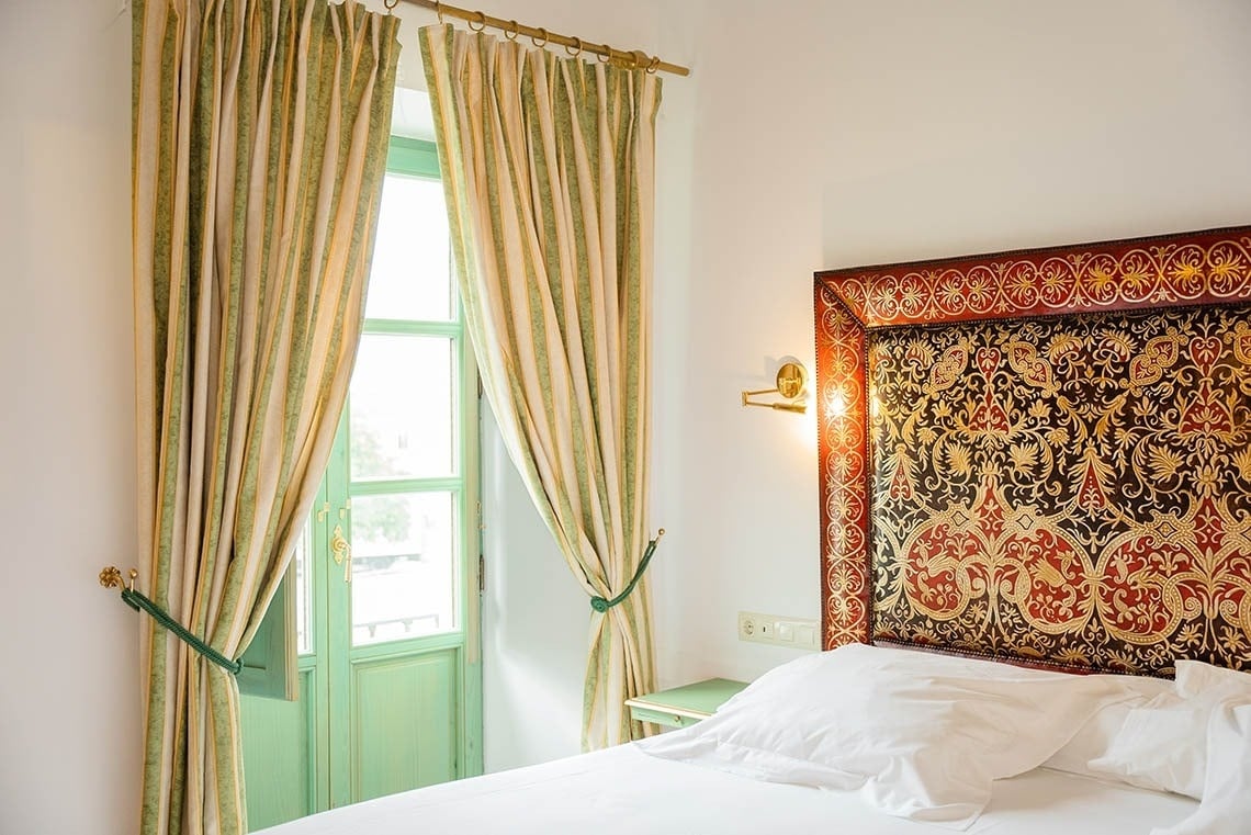 Un dormitorio luminoso presenta una cama con un cabecero ricamente decorado en rojo y dorado, flanqueado por cortinas a rayas verdes y crema que enmarcan una puerta-ventana de un suave tono verde.