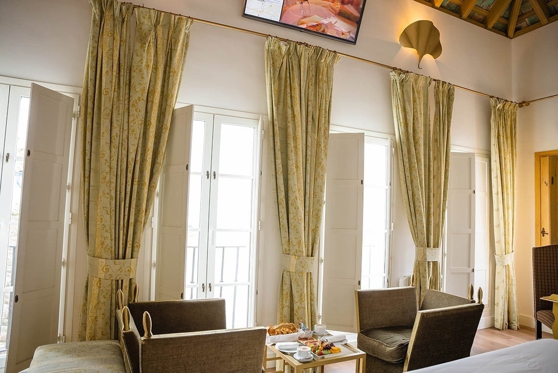 Una luminosa habitación de hotel presenta grandes ventanales con persianas blancas y cortinas con estampado dorado, un televisor en la parte superior y un desayuno servido en una mesa entre dos sillones.