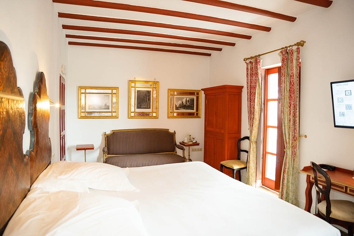 Un dormitorio de estilo cálido presenta una cama grande, vigas de madera expuestas en el techo, un armario rojo y obras de arte enmarcadas en dorado.