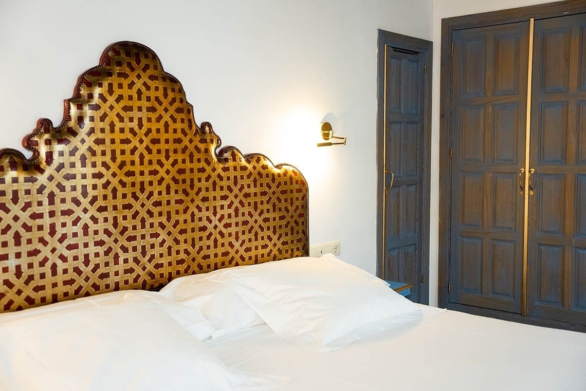 Una cama con un cabecero ornamentado de patrón geométrico dorado y rojo domina la escena, flanqueada por una lámpara de pared y un armario de madera oscura con detalles dorados.