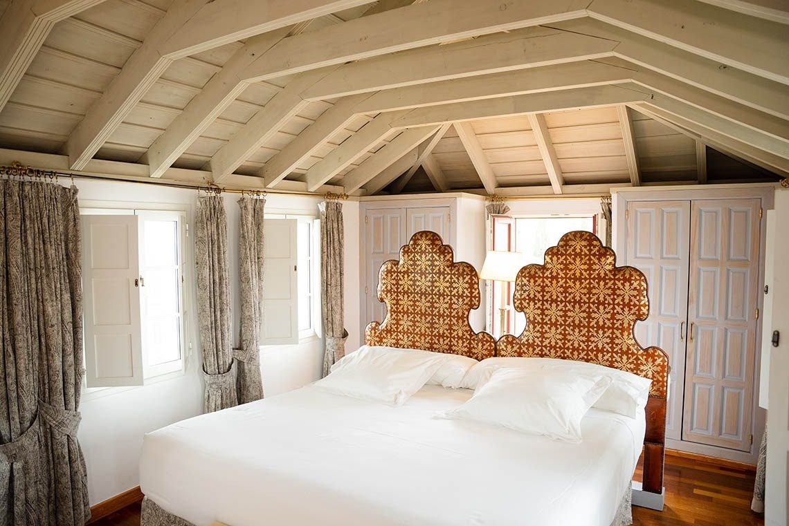 Un dormitorio acogedor presenta una cama con dos cabeceros ricamente estampados, cortinas claras enmarcando ventanas, y un techo abuhardillado con vigas de madera expuestas.