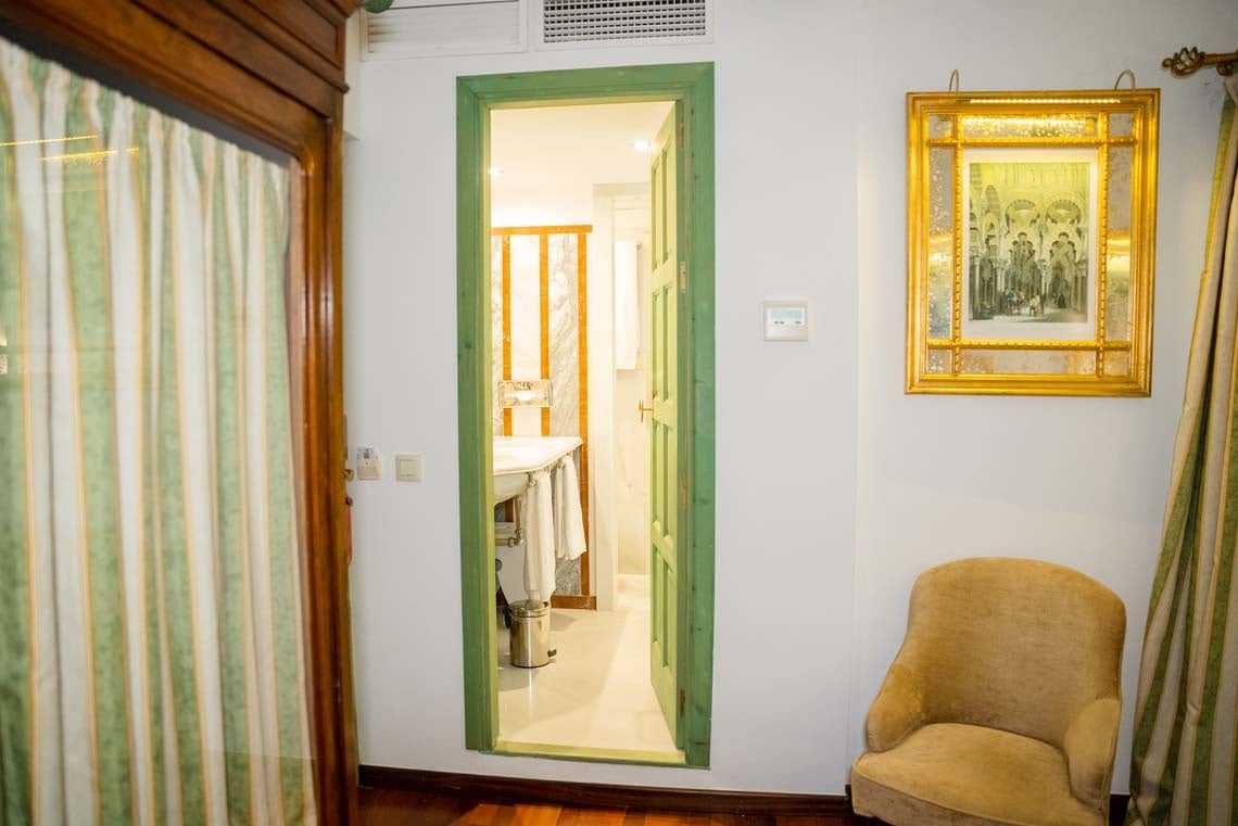 Charming hotel room with view into bathroom, featuring artwork and armchair, located in Juderia: Cordoba.