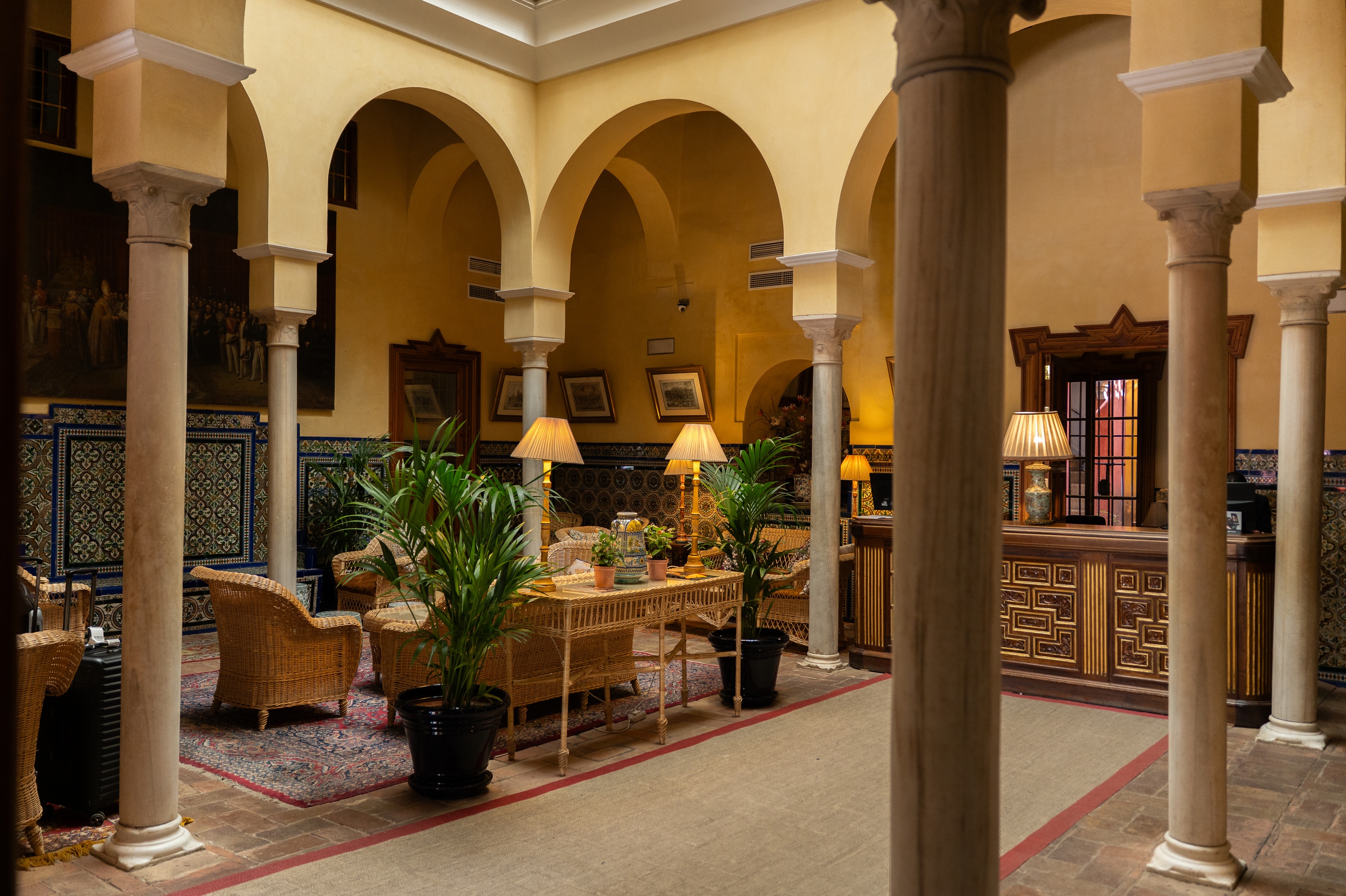 Patio en Judería Sevilla: Lámparas, plantas y sillones de mimbre bajo arcos.