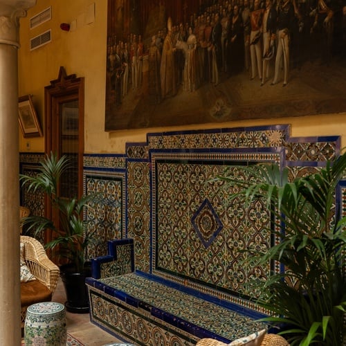 Courtyard seating area in Juderia: Sevilla, featuring ornate tiles, a large mural, and wicker furniture.