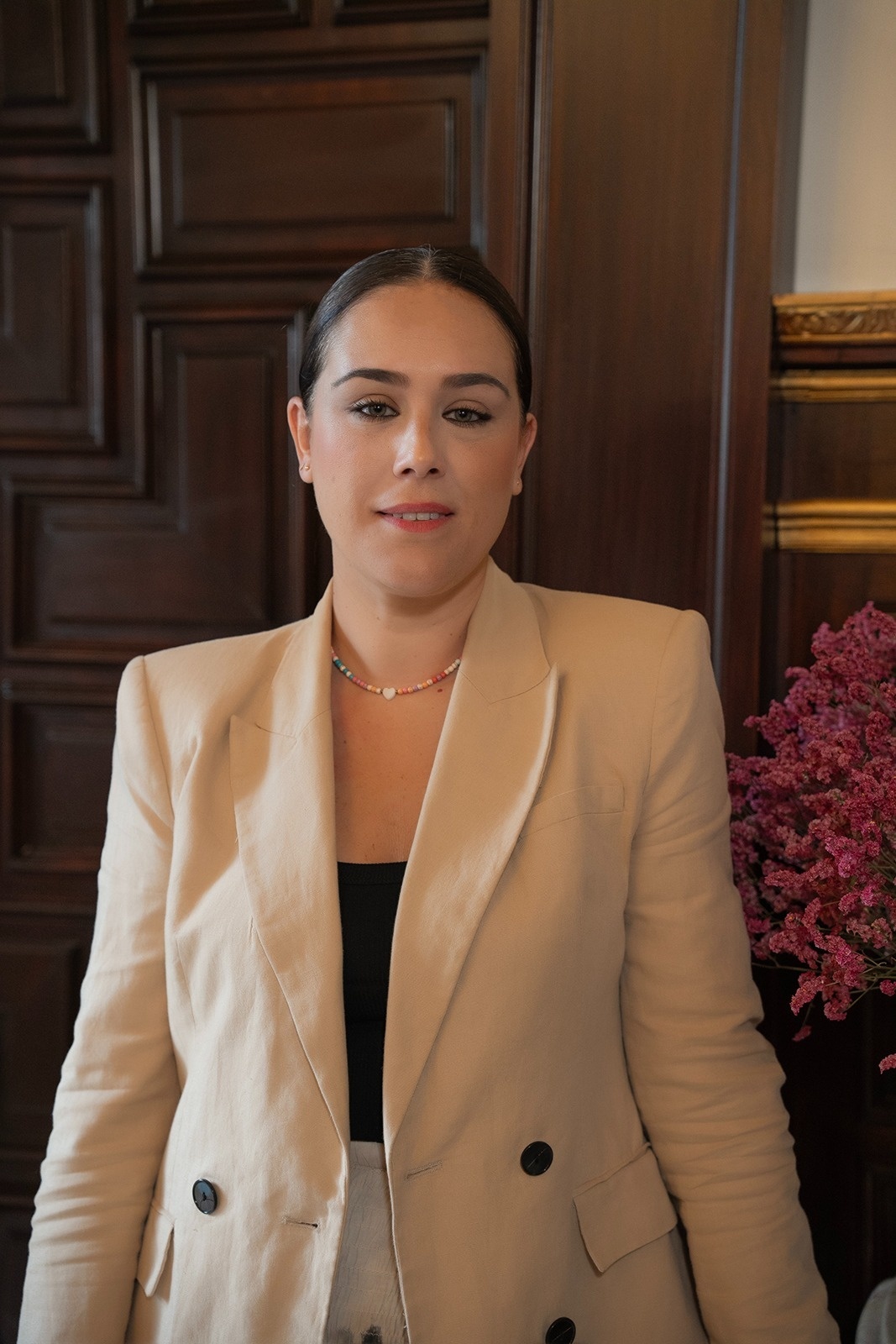  a woman wearing a blazer and a necklace stands in front of a wooden door 
