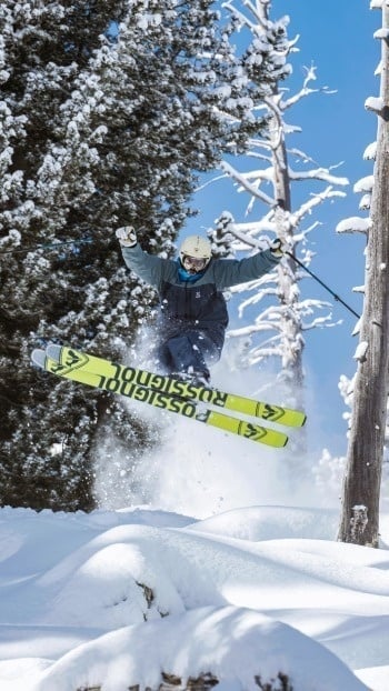 Un skieur en combinaison de ski et casque jaune est en plein saut, avec ses skis jaunes, au milieu d'un paysage enneigé parsemé d'arbres couverts de neige, sous un ciel bleu.