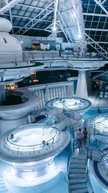 L'image présente un vaste complexe aquatique intérieur moderne et lumineux, avec plusieurs bassins étagés et des personnes se relaxant sous une grande structure de toit.