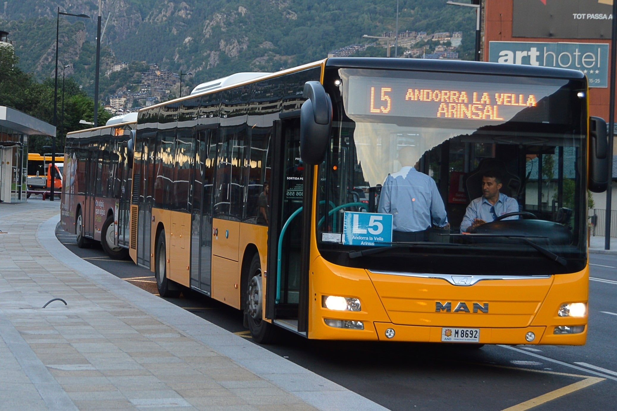 An orange articulated MAN bus with "L5 Andorra La Vella Arinsal" displayed on its front is shown in a city setting, with mountains and buildings in the background.