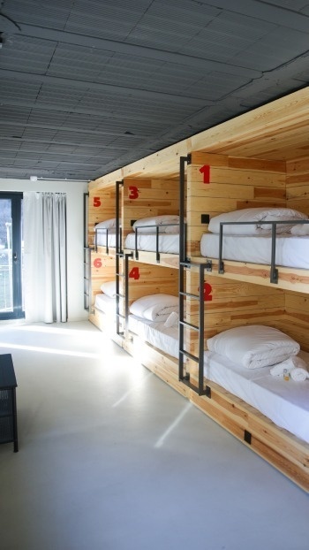 Cette image représente une chambre de style auberge de jeunesse avec plusieurs lits superposés en bois numérotés en rouge, des draps blancs, un plafond gris foncé et une fenêtre avec des rideaux blancs à gauche.