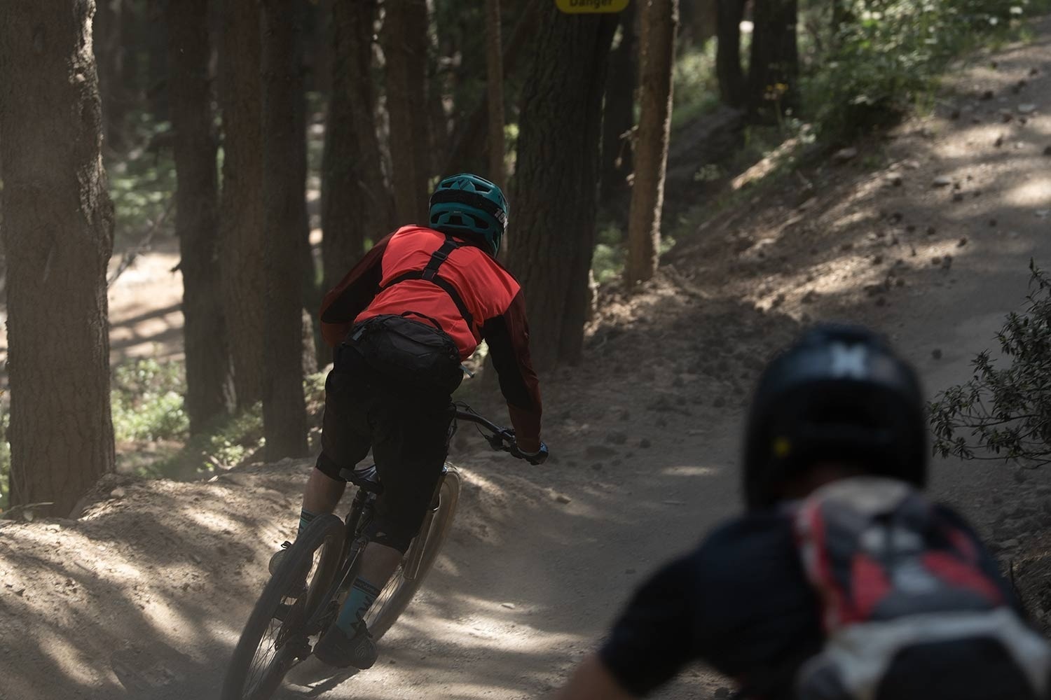 A mountain biker in a red shirt and teal helmet leans into a dusty turn on a sun-dappled forest trail, with another biker blurred in the foreground.