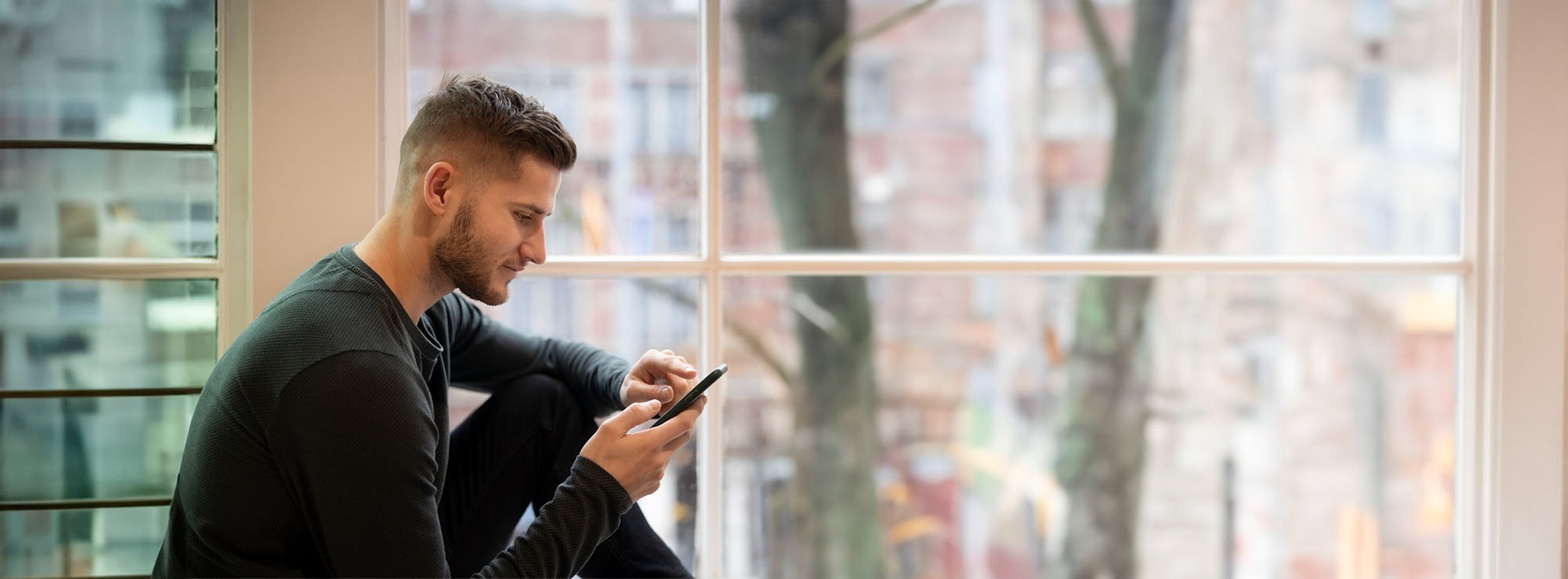 un hombre se sienta en el alféizar de una ventana mirando su teléfono celular