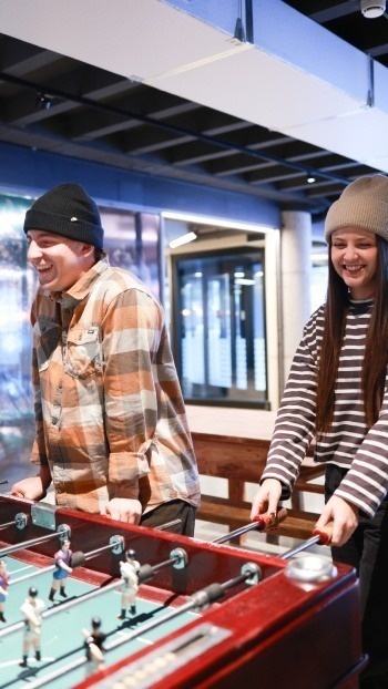 A man and a woman wearing beanies and casual attire are happily playing foosball.