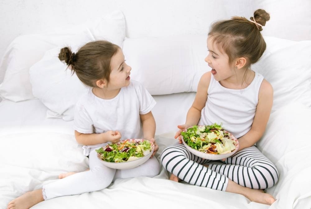 Zwei kleine Mädchen sitzen auf einem Bett und essen Salat