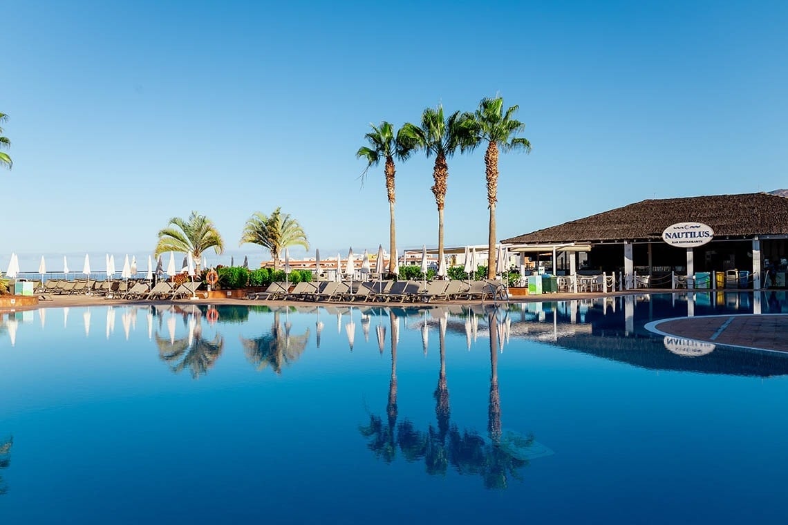La imagen muestra una piscina azul con palmeras y un bar-restaurante "Nautilus" con techo de paja, reflejados en el agua bajo un cielo despejado.