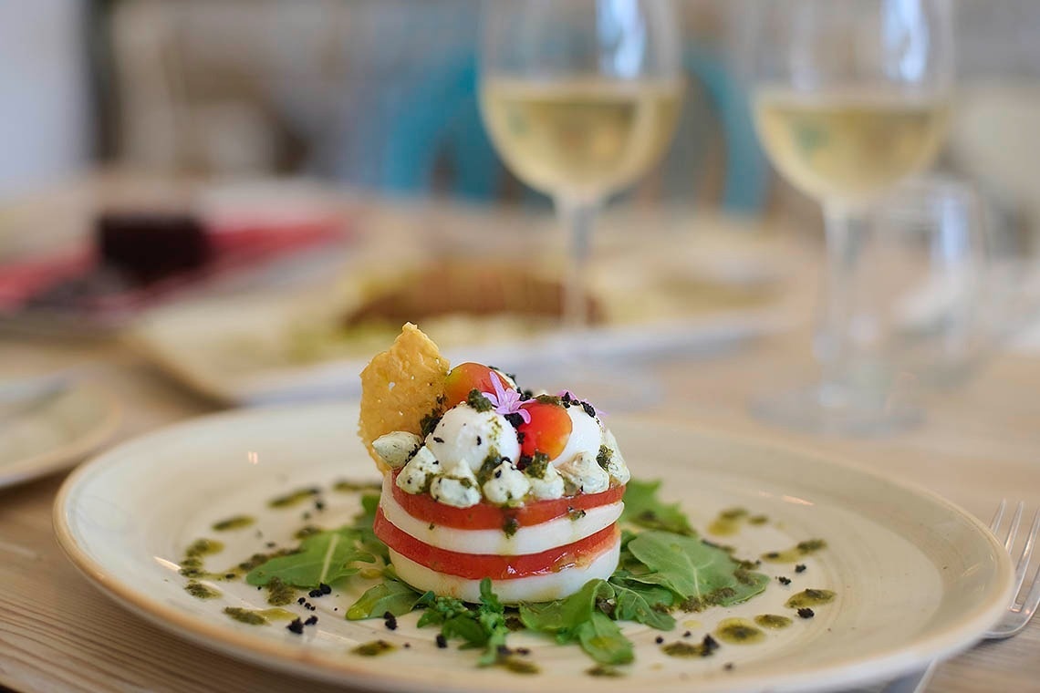 A gourmet Caprese salad, artfully stacked with mozzarella and tomatoes and garnished with herbs and a cracker, is presented on a plate with arugula and a green sauce, against a soft-focus background of wine glasses.