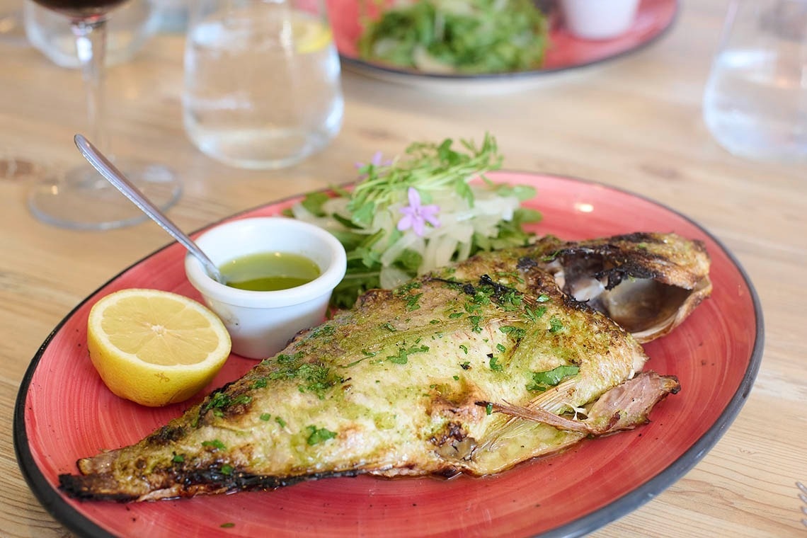 A whole grilled fish, seasoned with herbs and spices, is presented on a red plate with a lemon half, a side of green sauce, and a fresh salad, all set on a wooden table with drinks in the blurred background.