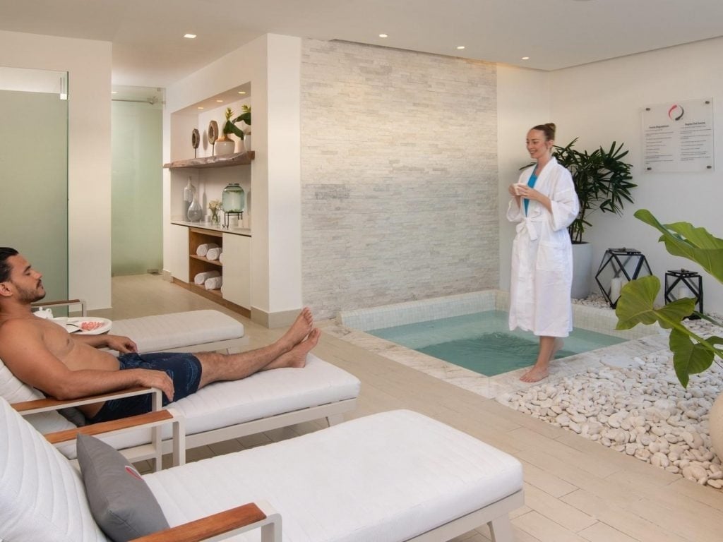 a woman in a bathrobe stands next to a man in lounge chairs in a spa
